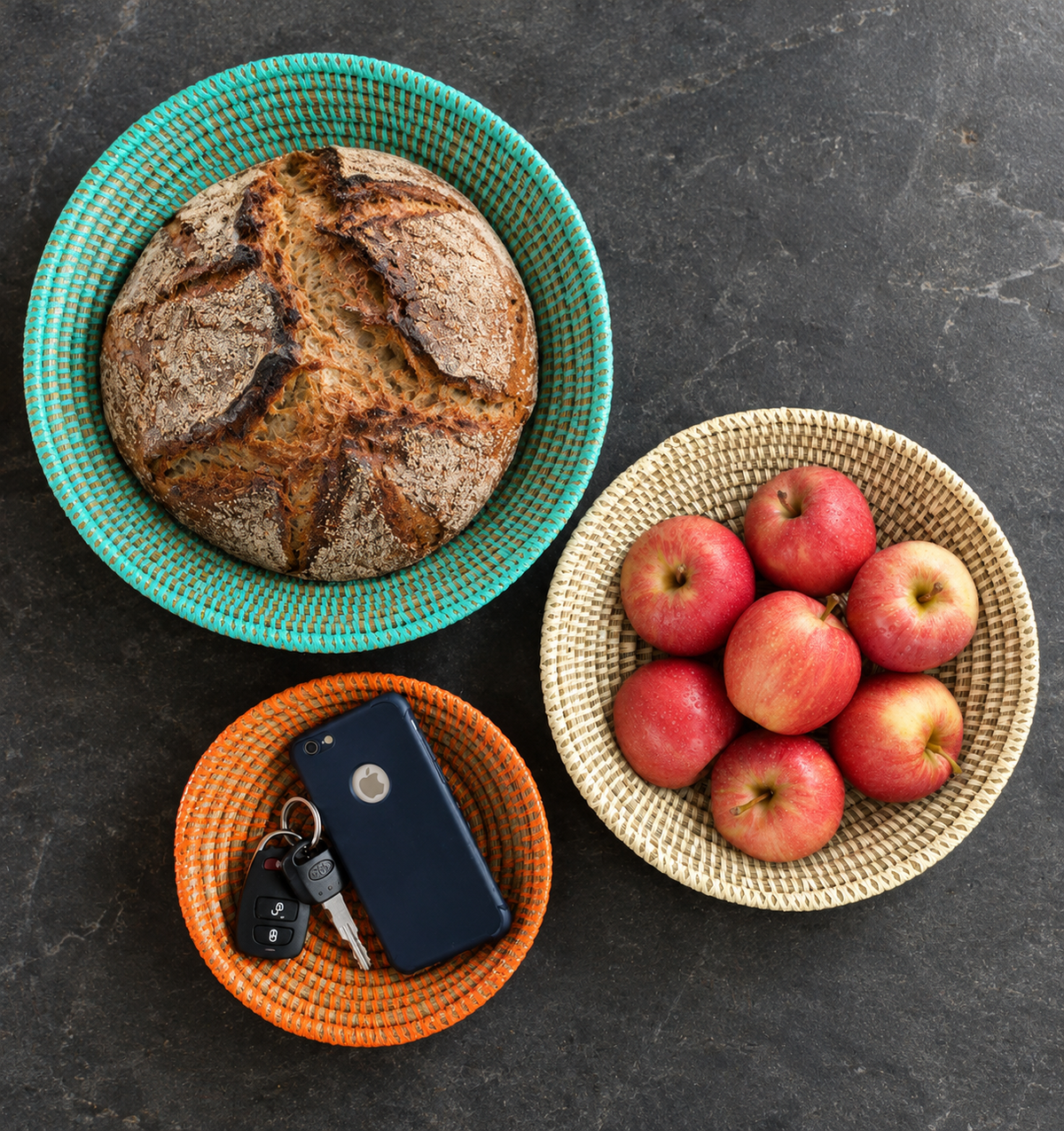 Three woven round bowls arranged on a dark stone surface, styled with a loaf of bread in a turquoise bowl, red apples in a cream bowl, and keys in an orange bowl.