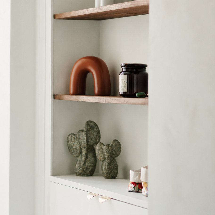 A built-in shelving unit with three rustic wooden floating shelves and a white lower drawer, styled with a white vase, a terracotta arch sculpture, a dark amber glass jar, and two green cactus figurines against a textured off-white wall.